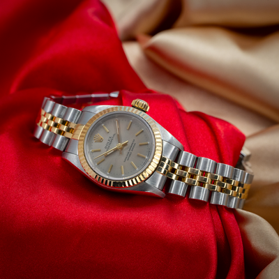 Rolex Oyster Perpetual Lady 67193 dial and two-tone bracelet with golden crown detail, on a red and gold satin background.