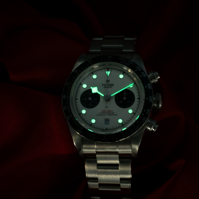 Tudor Black Bay Chrono M79360N-0002 dial with glowing hands and hour markers against a dark red background.
