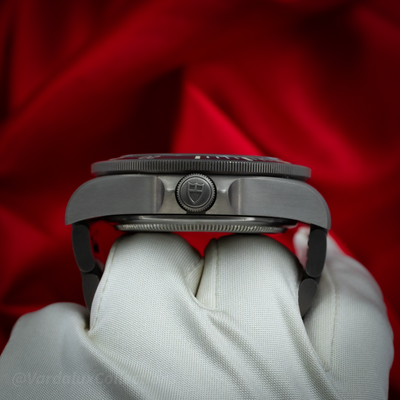 Tudor Pelagos 25600TN watch, left side case and crown detail, held by gloved hands against red.