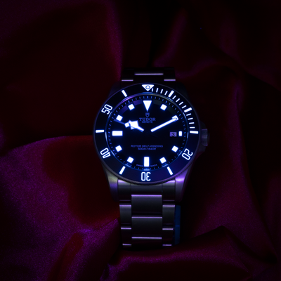 Tudor Pelagos 25500TN dial with glowing blue lume on markers and hands against a dark background.